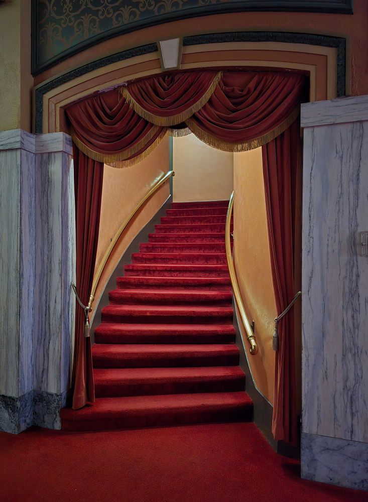 A plush, red carpeted stairway, curving gently to the right, with gold-trimmed, red theatre curtains decorating the entranceway.