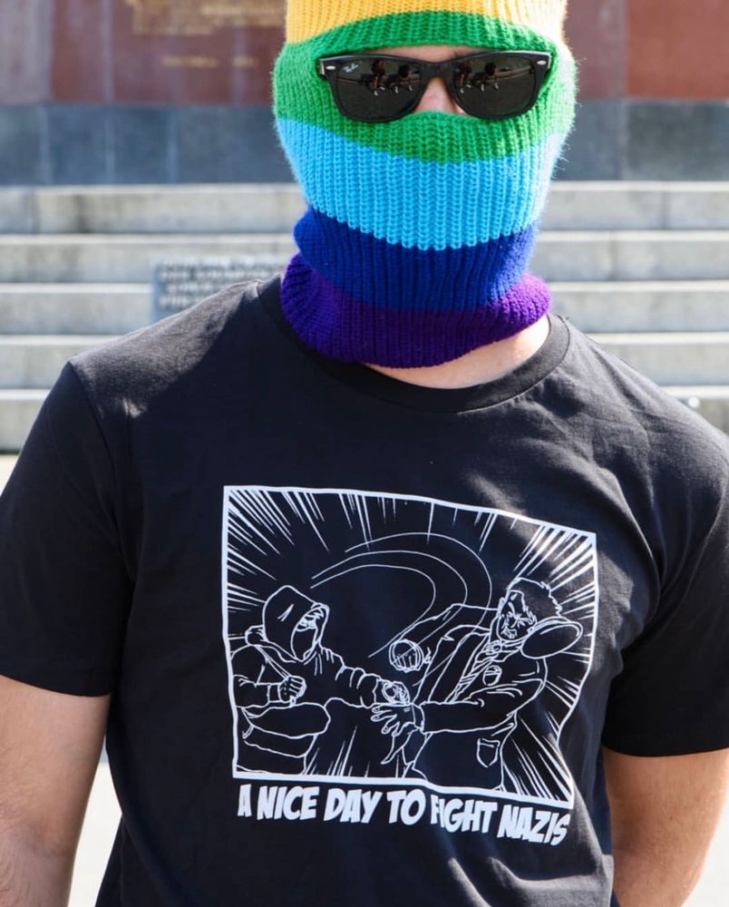 Auf dem Foto ist eine Person mit Regenbogen-Sturmhaube zu sehen, die ein schwarzes T-Shirt mit der Aufschrift "A nice day to fight nazis" trägt. Das Motiv über dem Schriftzug ist im Comic-Stil: Eine vermummte Person schlägt einen Burschenschafter dem die Kopfbedeckung vom Kopf fällt.