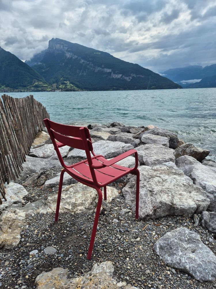 An einem See steht im Vordergrund ein roter Plastik-Sessel, daneben große Steine. Im Hintergrund ist eine Berglandschaft.