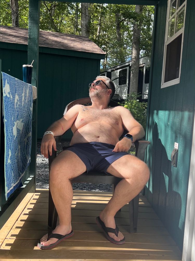 Picture of me, lounging reclined in a plastic chair, sitting on the porch of a cabin. Afternoon sunshine is dappled over my face and body. Wearing blue swim shorts, sunglasses and flip flops. 