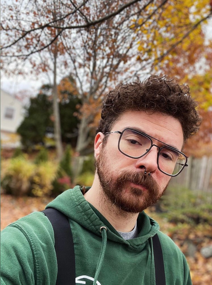 Selfie of Connor outside with autumn trees behind him, he’s in a green hoodie and overalls. Gay scruff beard mustache queer alt 