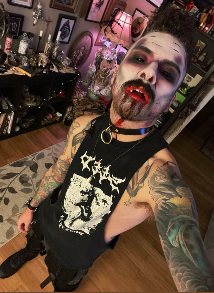 Connor in white face makeup, dark eyes and blood in his mouth. He’s in a black tank and combat boots. Tattoos queer gay goth alt goth makeup 