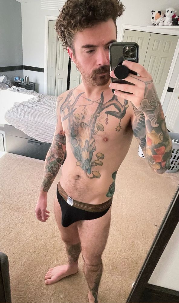 Full body Connor in a mirror selfie wearing a black thong. He has multiple color tattoos and a mustache. Gay queer hairy beard scruff bulge underwear 