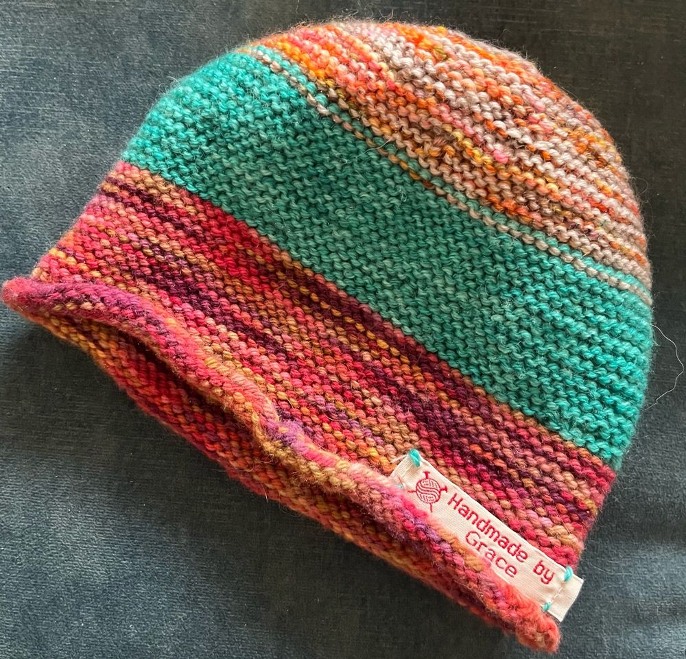 a photo of a colourful striped knitted baby hat in orange, grey, light blue, purple and pink, with a label sewed on the outer rim of the hat that reads 'Handmade by Grace'