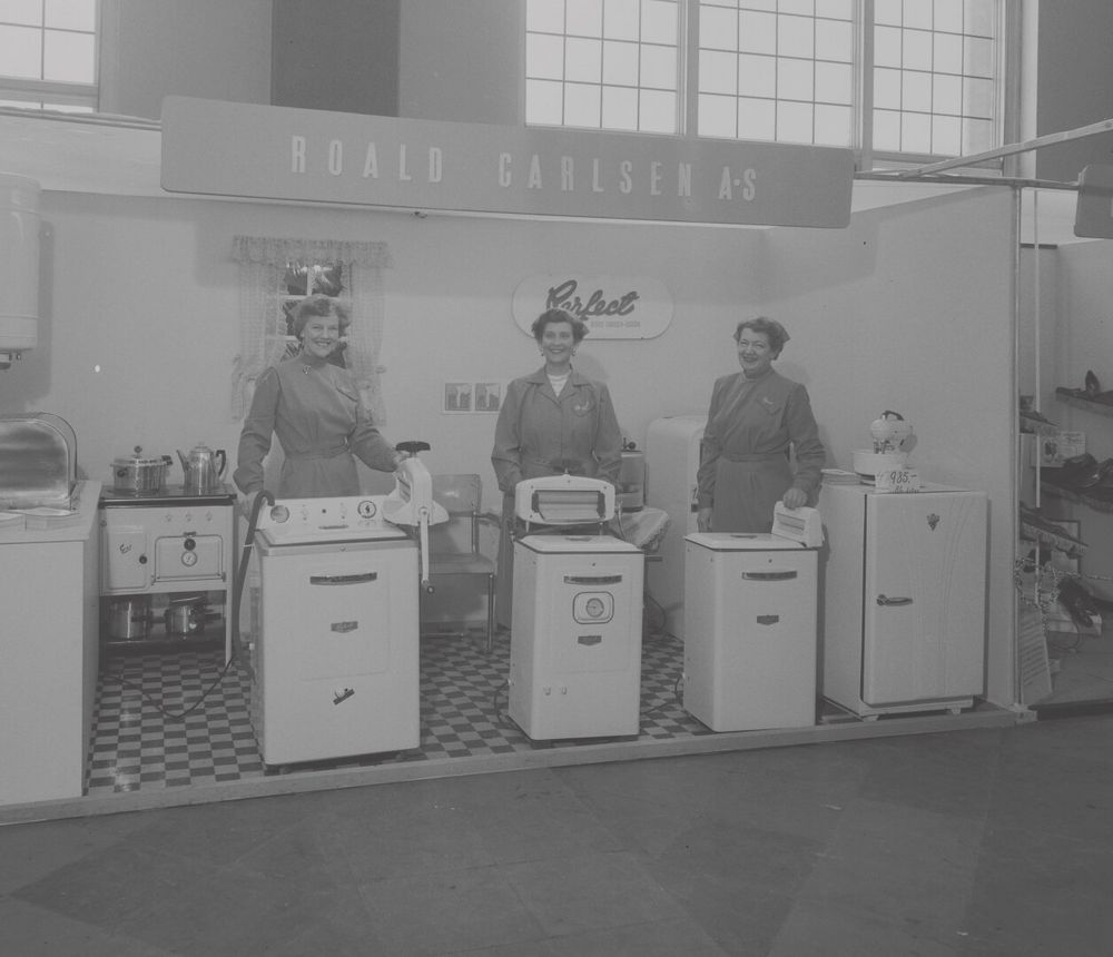 Three women standing in a trade booth under the sign "Roald Carlsen A/S", with washing machines and fridges and other appliances.