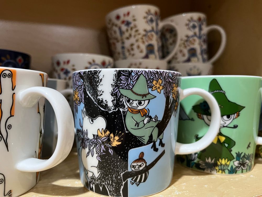 Photo of Moonin mugs - to the left you can kinda see the Hattifatterners in an orange background, Snufkin, Moomin and Little Mai in a tree in the middle and a Snufkin mug with a green background to the right. 
