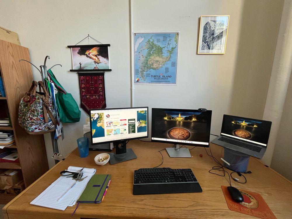 Photo of a desk with a computer, two screens and the decolonial atlas map on the wall surrounded by a print titled return the empire to the wind in which North America burns and another print titled halocline showing an iceberg in the shape of a heart melting surrounded by birds and fish