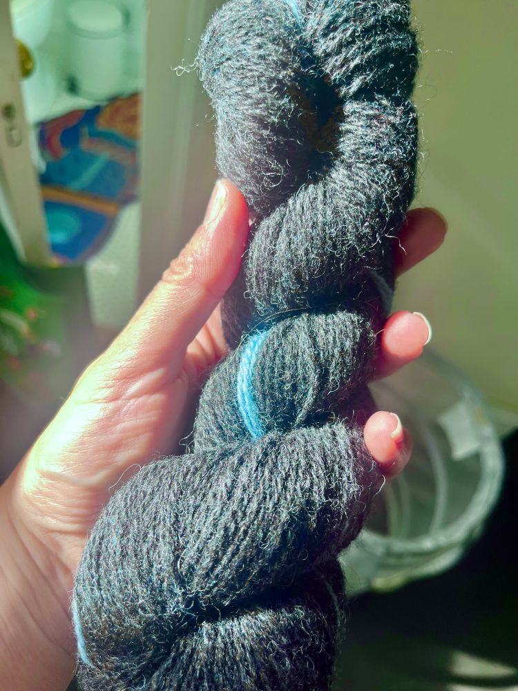 A hand holds a skein of woolen yarn of a dark charcoal gray color with a faint blue tint, the color called bloth in old Norse. It is held in bright sunlight which makes it look somewhat pale, the color of slate. 