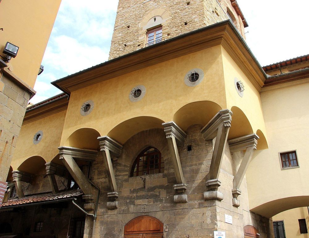 Photograph of a building in Florence. A tall thin stone section rises up, from street level several stories. About the level of the second story, a yellow section sticks out from the outside of it, awkwardly wrapping around the outside of the stone part, supported by elegant sticky-outy triangular struts. The yellow section has several small circular windows, much too small for a human to climb through, barely large enough for a chubby cat.