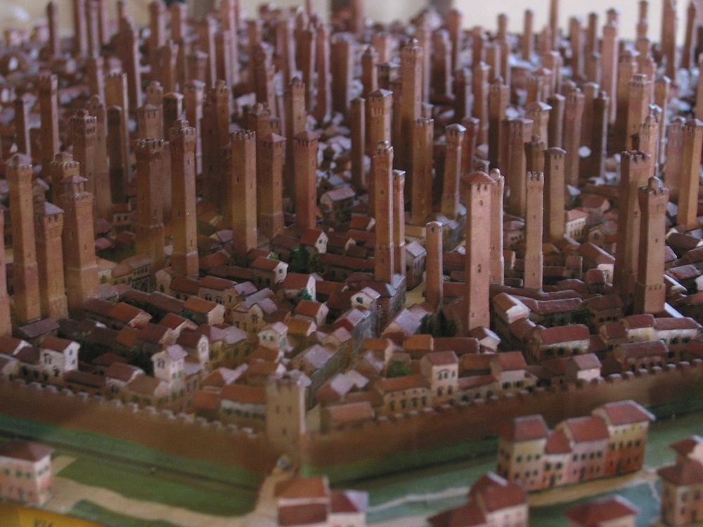 Photo of a model of the Medieval city of Bologna, with so many earthy pink tall skinny towers sticking up from every block of the terra-cotta-roofed town that it looks like plant seeds starting to come up in spring. Around the edge you can see the city's moat and battlemented walls, looking tiny compared to the towers which rise to six or seven times the height of the three-story buildings around them.