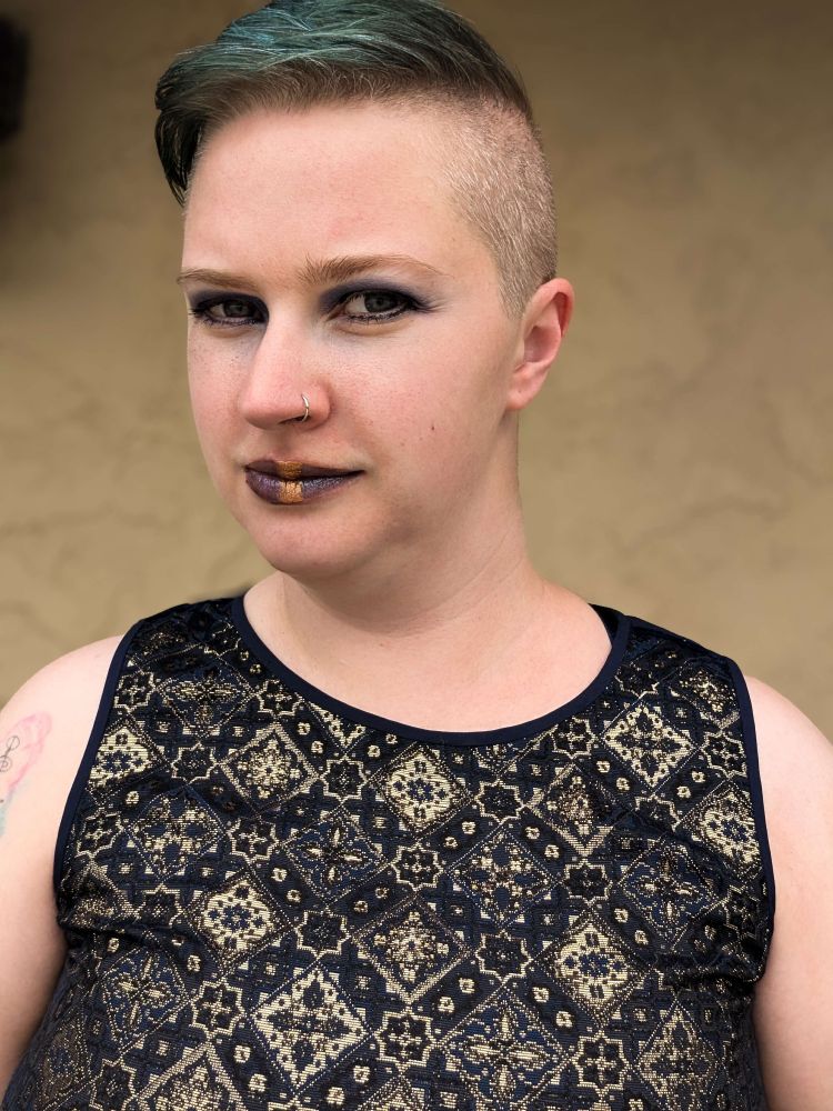 Mav (white, androgynous) is wearing a sleeveless navy top with an intricate gold pattern. Their makeup is dark, including blue lipstick with a gold strip down the middle. They look mysteriousand intense.