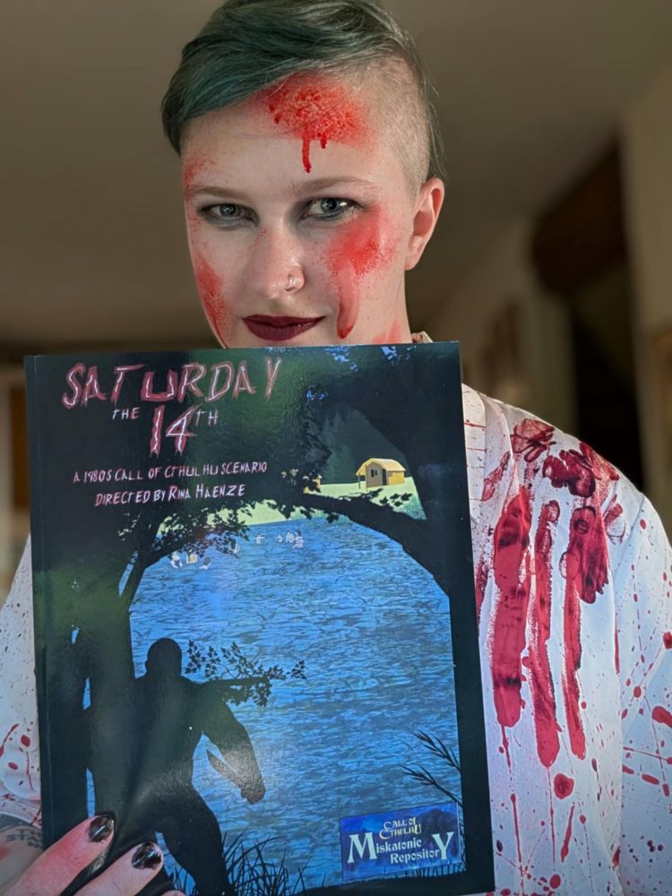 Rina (white, androgynous) is wearing a blood spatter shirt and is covered with bloody makeup. They're looking at you with a mysterious smirk. They're holding a copy of their scenario "Saturday the 14th", which has a mysterious shadowy figure holding a knife and watching people swimming on the cover. 