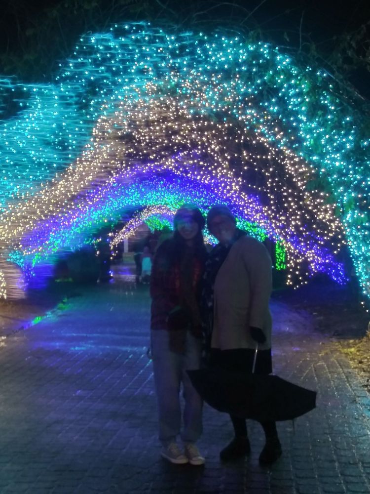 Two people smile for a picture in front of arches of bright colorful Christmas lights.