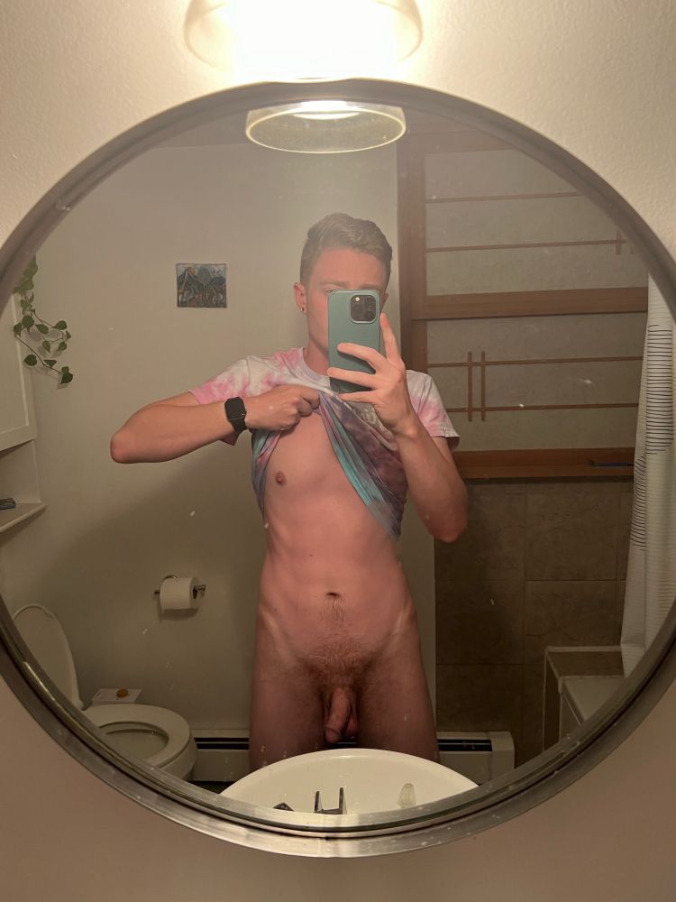 Ian standing mostly naked in front of a bathroom mirror, soft cock hanging while lifting one side of his tie-dye shirt to show a nipple