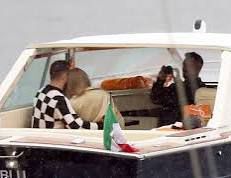 a paparazzi photo of the back of travis kelce in a black and white check shirt and taylor swift in a beige jacket heads. they are on a boat. someone else in shadow is taking a photo of them