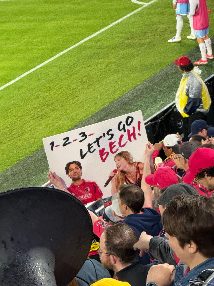 a fan holding a poster with a man and taylor swift on it that says “1-2-3 let’s go bech”