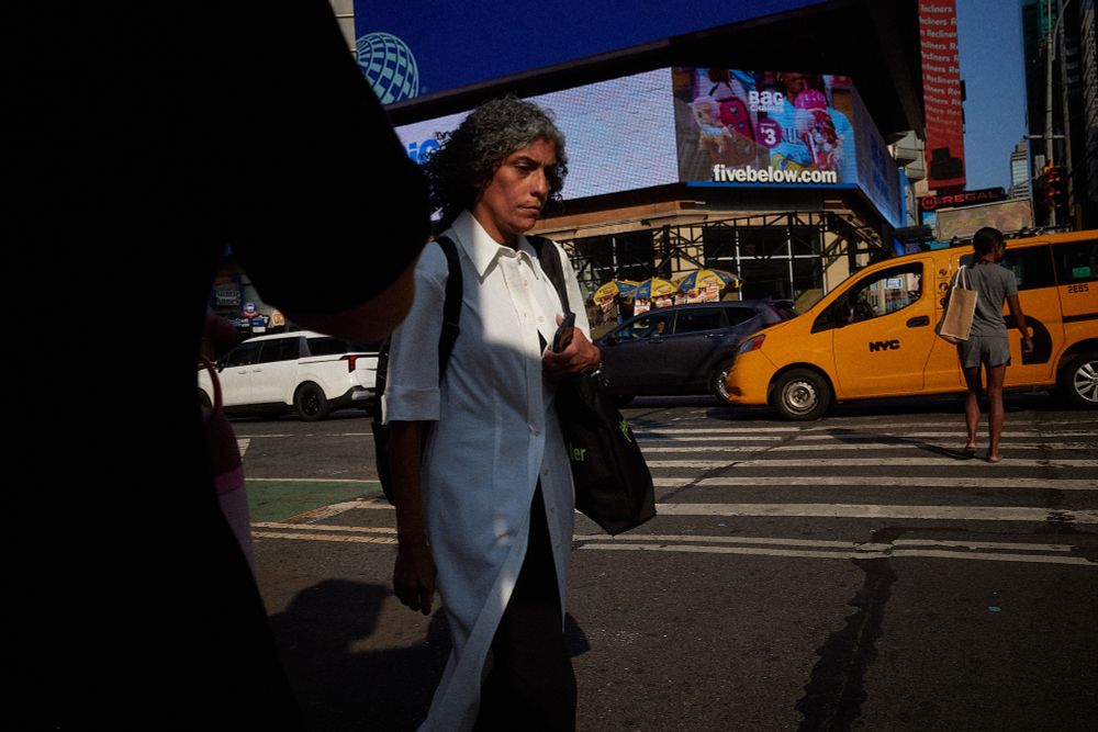 A woman walks through late afternoon light, a small square of it illuminating her face. Taxicabs and billboards fill the background. 