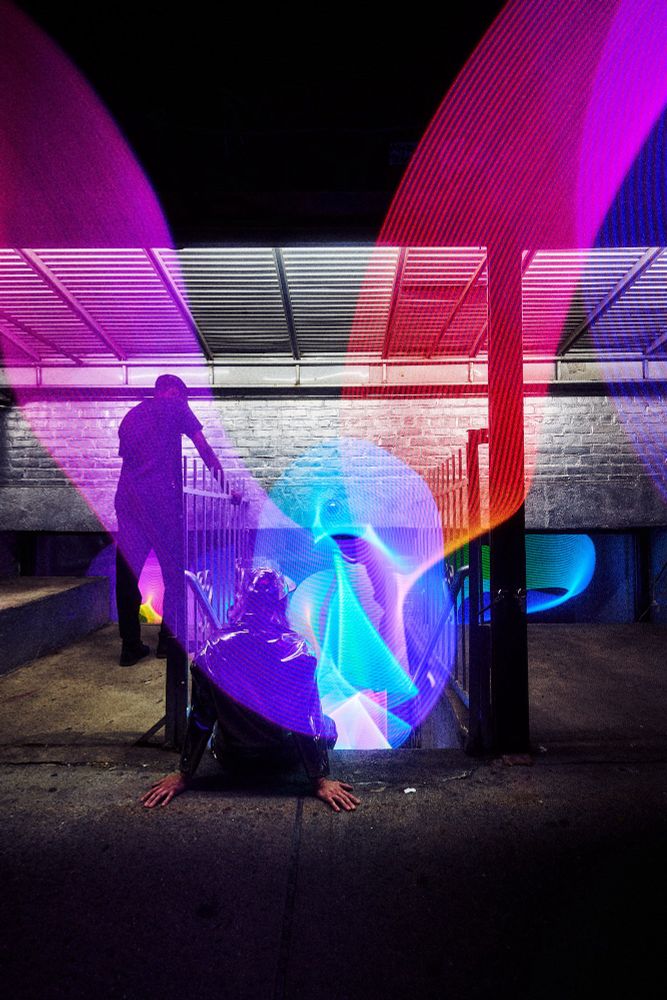Two figures from behind, one seated on a set of stairs leading to a basement gallery space, one standing and leaning on a fence above the stairs, with multicolored ribbons of light flowing past them and into the gallery space.