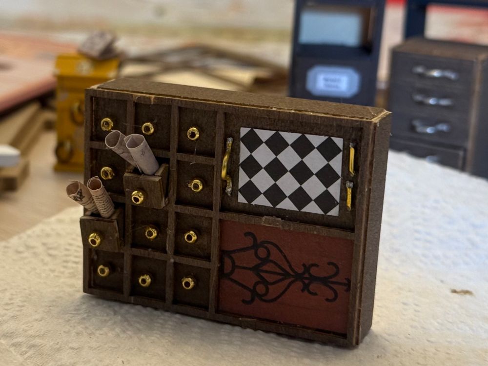 a wide dark brown cabinet with a 3x3 grid of square little drawers with golden bead handles on the left. two of them are open with little curled up scrolls of paper sticking out. the right side has like. a black and white diamond pattern in a rectangle on top with some golden handles glued on either side of it. the bottom has a fancy black symbol on the wood. in general it's a little rough and fucked up looking but it's cute