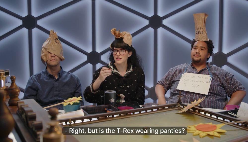 zac, emily, and lou at the table. all three have paper bags on top of their heads in some way - zac has managed to split his in a way that covers the top half of his face like an emo helmet, emily's is crumpled on top of her head like a tiara made of garbage, and lou's is somehow standing completely vertical upside down in a way that shows that the bag has the word FARTS on the front in big black marker. lou also has a sign around his neck over his shirt that says "Do not feed me aioli no matter what I say". emily with a straight face asks "Right, but is the T-Rex wearing panties?"