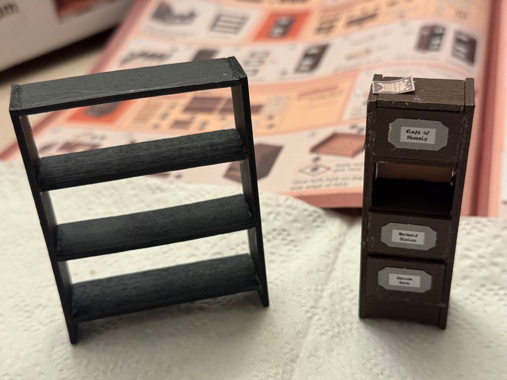 a very basic black wooden standing shelf (left) and a more complicated dark brown cabinet with little signs that list magical ingredients on the drawers (right) with the instructions booklet in the background. the right cabinet has glue strands all over because i am just learning how to do this