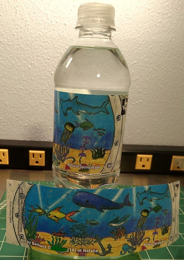 Two copies of a water-bottle label, one wrapped around a bottle and one removed, spread open to show the full label. The full label is a drawing of sea creatures as seen in blue water through a round porthole. The sea creatures include a whale, hammerhead shark, jellyfish, octopus, fish, seaweed. An anchor is partly buried in yellow sand. 