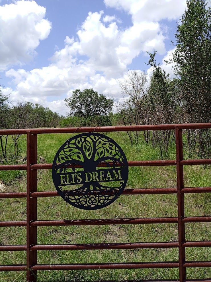 On a red metal gate, a circular black sign contains the text ELI'S DREAM across a cutout design of a large tree with branches spreading as far above as its roots spread below. Behind the gate are yellow&green grass, brown&green trees, blue&white sky. 