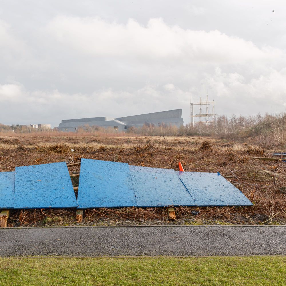 A traffic cone peaks out from beneath flattened blue hoarding with wasteground beyond. On the horizon is the Riverside Museum with the Science Centre Tower and Tall Ship beside it. 