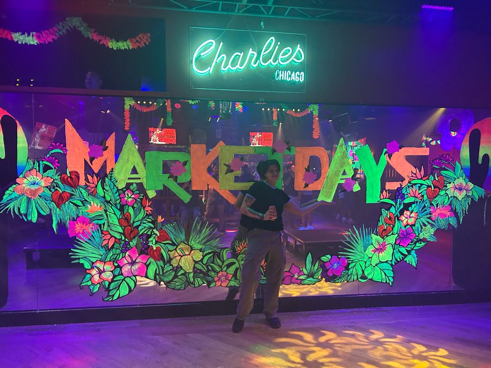 Micki standing infront of a mirror wall at Charlie’s. There is a neon floral mural painted on the glass that says market days