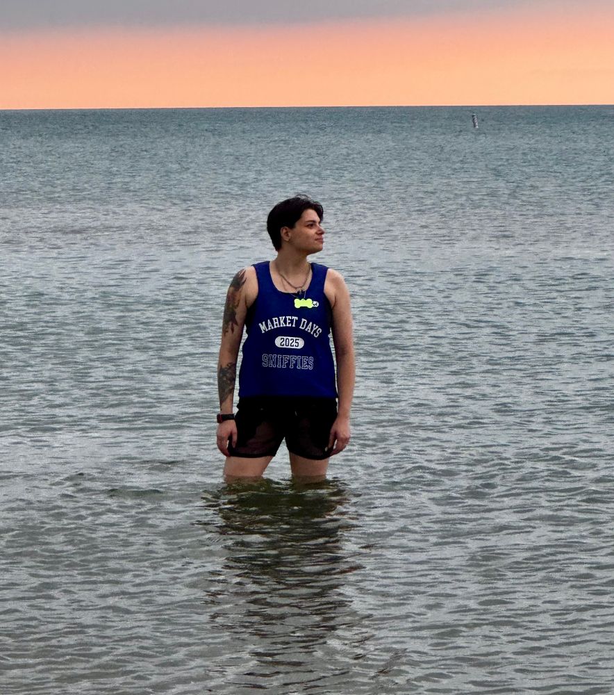 Micki wearing a blue Sniffies brand Market Days jersey while standing thigh deep in Lake Michigan during sunrise 