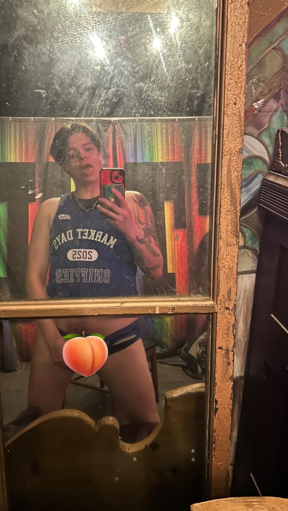 Micki posing in a mirror, tongue out, pulling their briefs down. Crotch is hidden by a peach emoji 