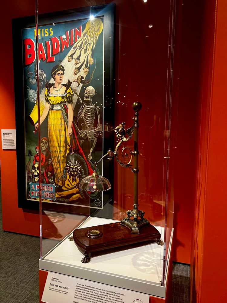 A spirit bell encased in glass sits in front of a tall lithograph advertisement for Miss Baldwin.