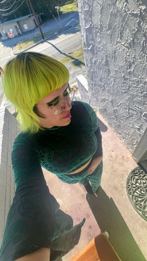 a trans man standing on his front porch with bright green hair. he is wearing a flowy green and black two piece set with design such has a pentagram on the front. 