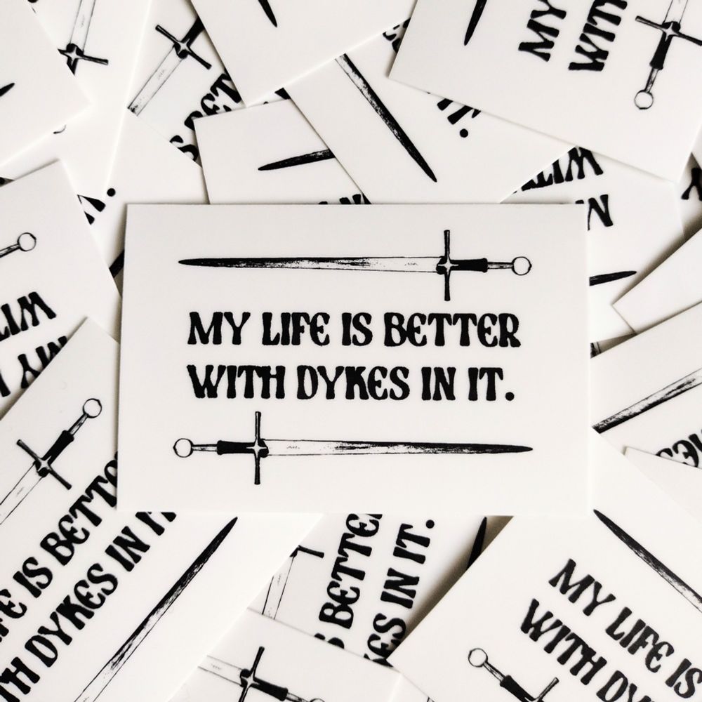 a black and white sticker that reads "my life is better with dykes in it". above and below the text are two swords 