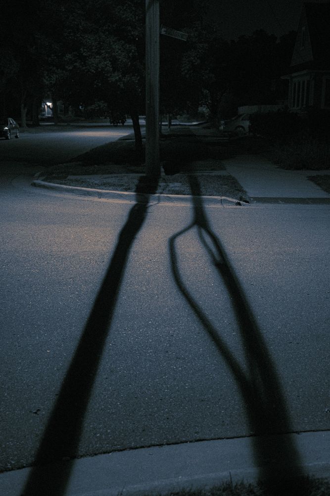 Across the street a shadow grows from a utility pole on the other side of the road. Another, parallel to it, comes from an unknown source. 