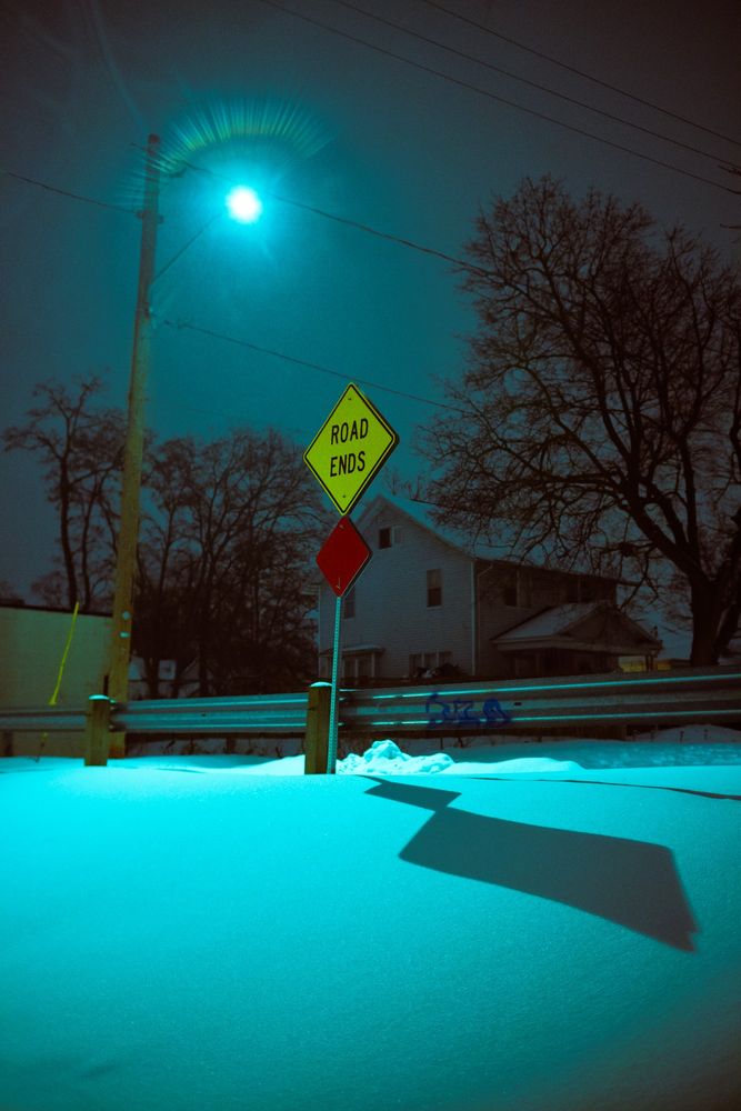 A street sign on the crest of a small snowy hill with the words "ROAD ENDS". A street light stands in the background, projecting a shadow of the sign on the snow. 