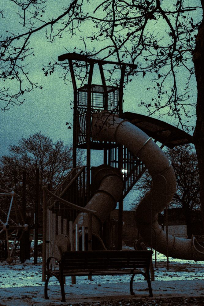 A jungle gym and slide in a playground. The sky is dark and overcast. Snow dusts the ground. There are no children. 