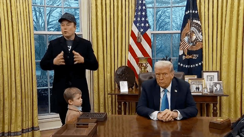 A scene in an office resembling the Oval Office. A man stands, gesturing with his hands, wearing a black coat and a hat. A young child stands beside him, looking towards the man. In the background, a man with light-colored hair is seated at a desk, wearing a dark suit and a blue tie, looking serious. The walls are adorned with yellow curtains and two flags.
