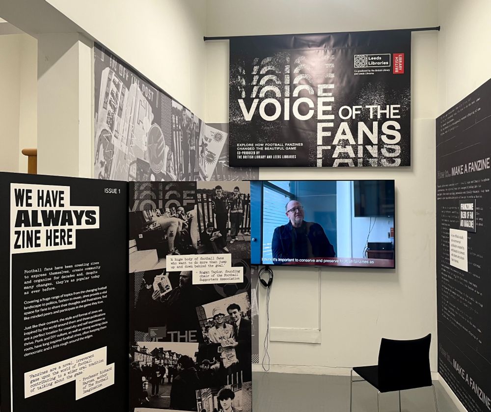 The entrance to the exhibition. There is a black cardboard display to the left of the image detailing the history of zine culture and starts with the title 'We have always zine here'. In the background is a black and white banner that reads 'Voice of the fans' with a video playing beneath it with a chair for visitors to sit on.