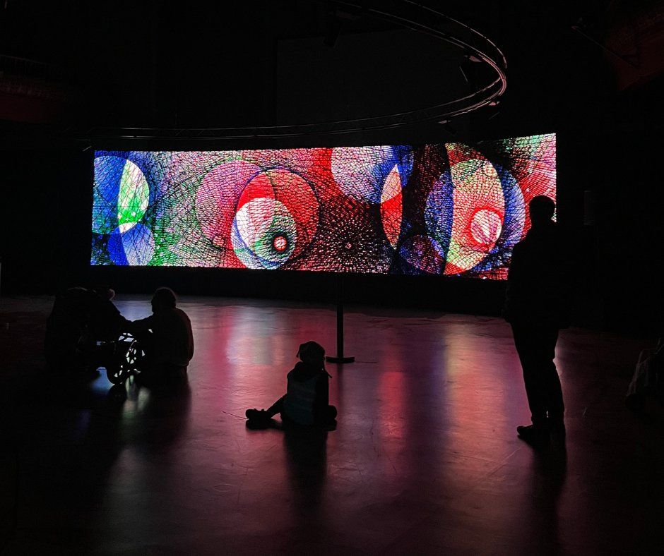 Spirograph Reimagined playing in Leeds City Museum as audience members sit and stand in front of it. The colours in the scene captured are bright tones of blue, green and red and resemble shapes you are able to create with a spirograph.