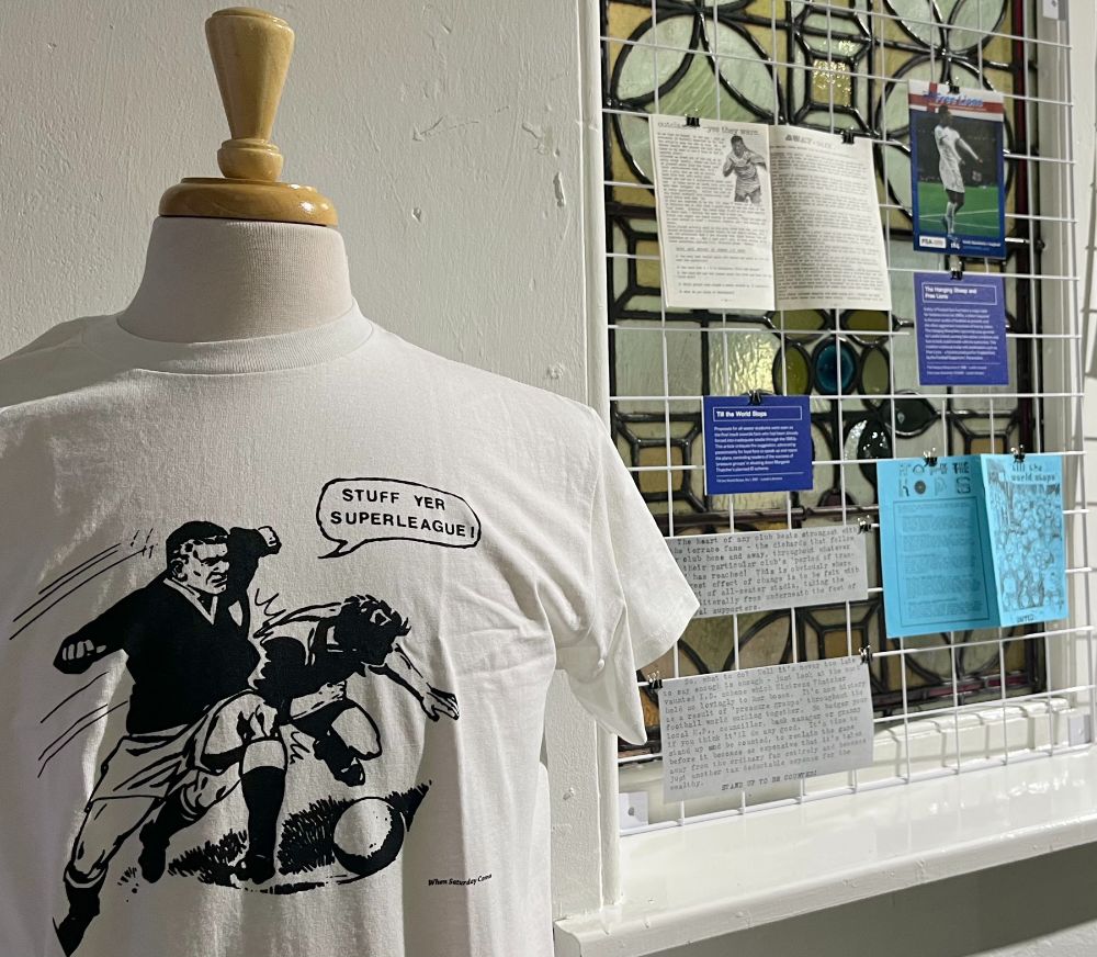 A t shirt displayed as part of the exhibition, its base is white and has a black illustration of a tackle in which one player is saying 'Stuff yer superleague' to the other. To the right of the t shirt in the background are a few zines and handouts displayed on a wire frame.