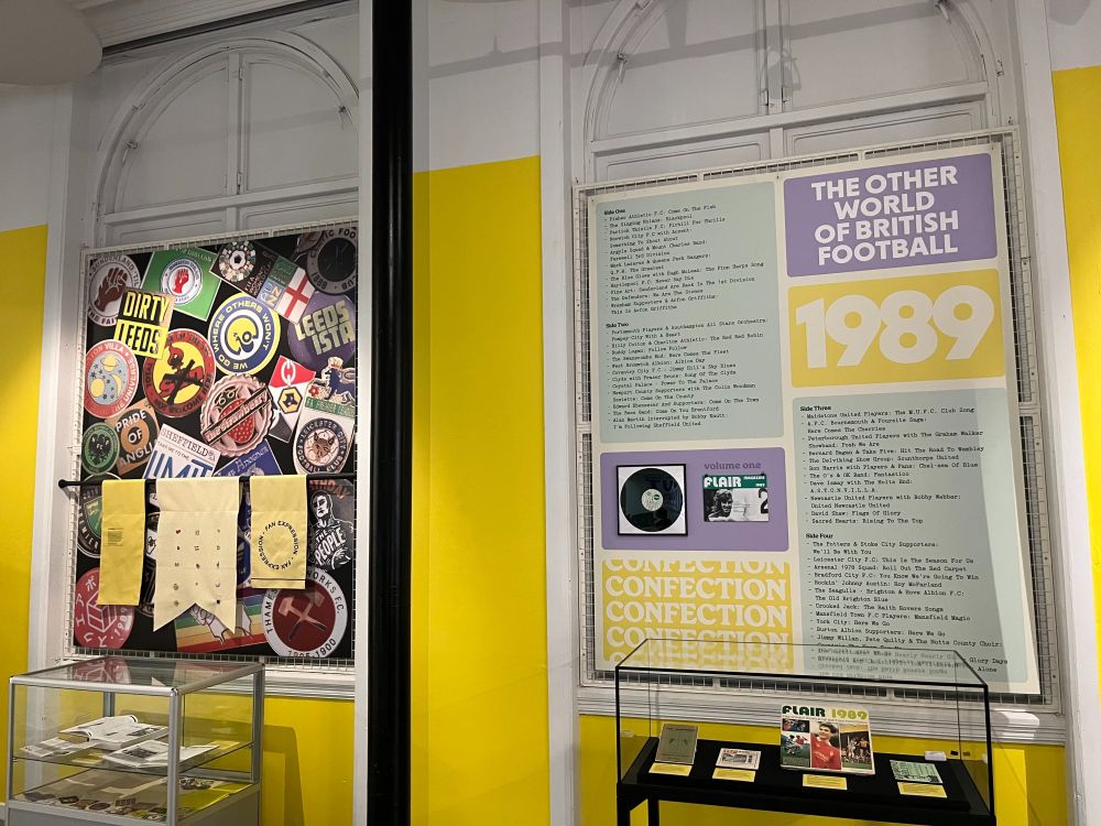 A section of the exhibition capturing football memorabilia such as badges, zines, and stickers. These items are depicted in vinyl stickers and prints on the wall and also in display cabinets.