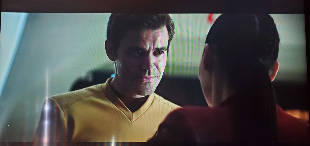 An unedited snapshot from a paused TV. On screen is a scene from the Star Trek Strange New Worlds episode Subspace Rhapsody, showing James Kirk, as played by Paul Wesley, looking down on La'An Noonien-Singh. The dead pixels appear slightly offset from both of his actual pupils, making him look like that rock monster sidekick Scotty had in the JJ Abrams movies