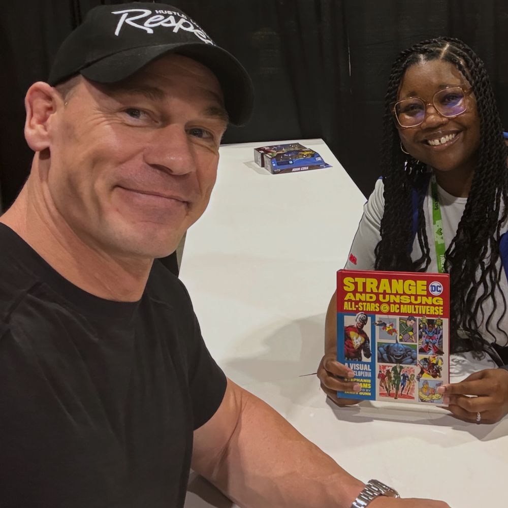 The image is of me holding a copy of Strange and Unsung All-Stars of the DC Multiverse at an empty John Cena table. 