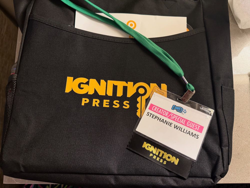 In the image is a black tote bag with the Ignition Press logo on it. My ComicsPro badge is sitting on top of it and says Creator/Special Guest Stephanie Williams and there is an Ignition Press ribbon attached to it. 