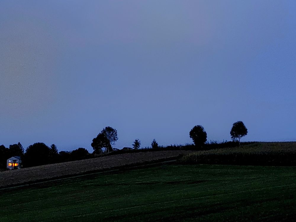 Wiesen und ein Feld am Abend. Am Horizont einzelne Bäume. Links ein abschüssiges Feld, am Bildrand ein Haus mit erleuchteten Fenstern. 