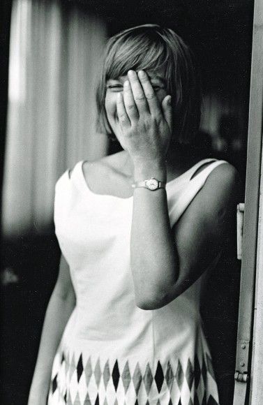 Schwarzweißportrait einer lachenden Ingeborg Bachmann in weißem Kleid. Sie hält die linke Hand vor ihr Gesicht. 