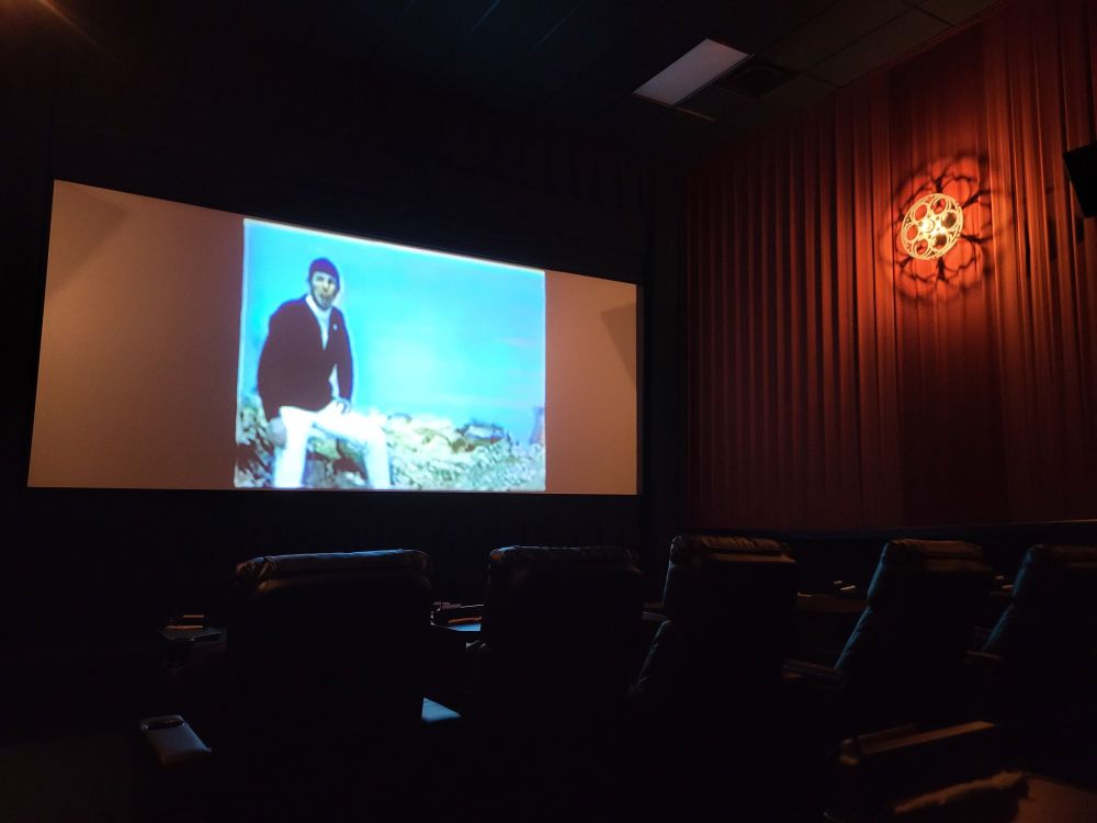 Dark empty Theater with a dim orange light coming from a fixture of a film reel on the wall. On the screen is Leonard Nimoy dressed in 70s fashion (jacket, turtleneck, white pants) sitting on a stony outgrowing. It is a film clip of him singing The Ballad of Bilbo Baggins.