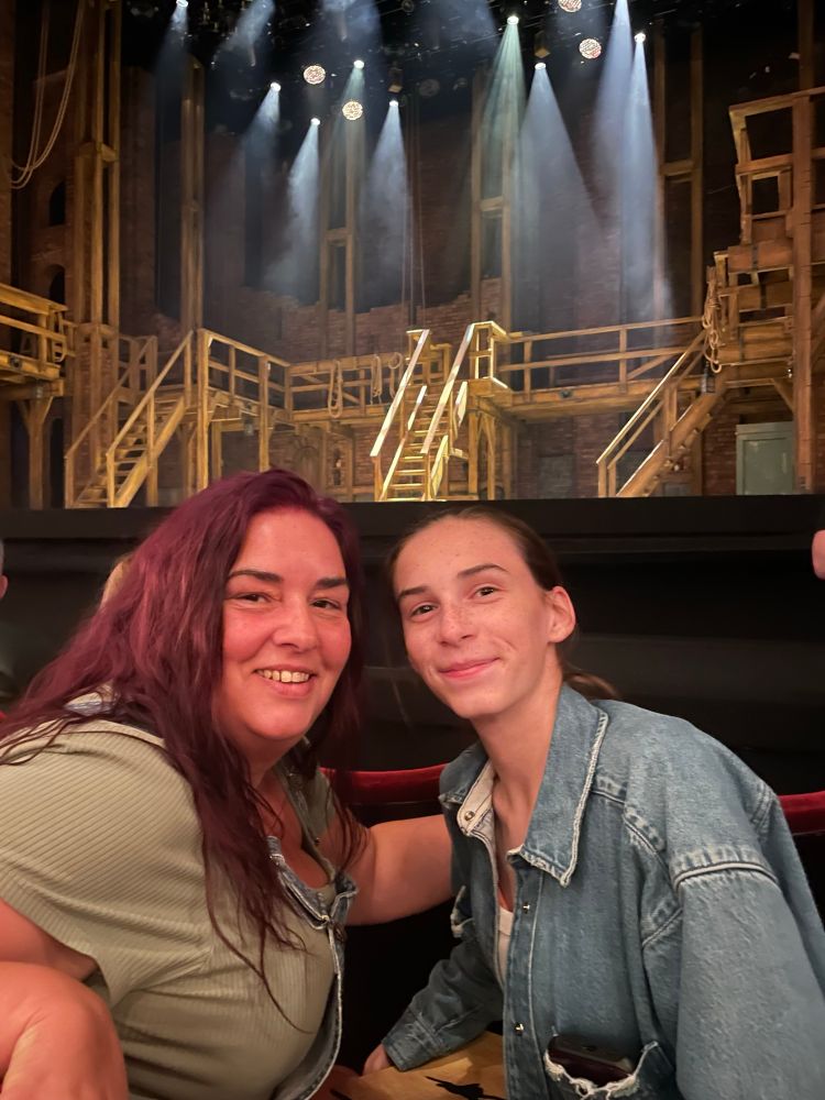 Stage of Hamilton in background Emma with purple hair. Wearing a green top. Emily hair back in denim. 