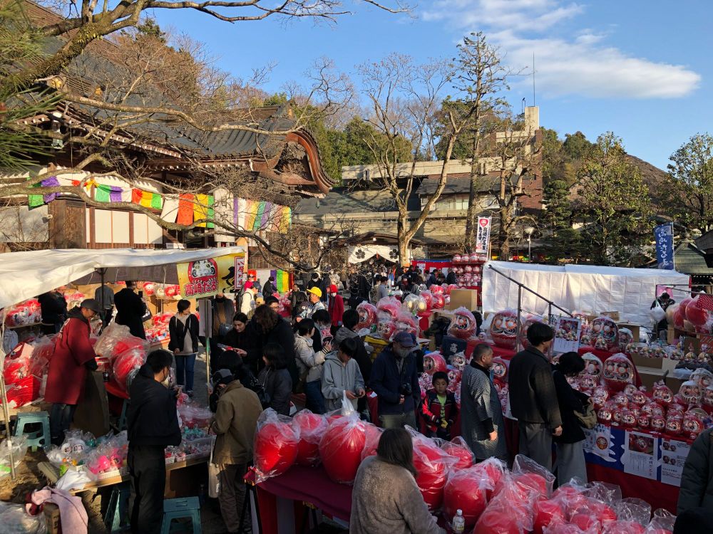 A view of Jindai-ji courtyard filled with stalls selling Daruma figures of all sizes, and crowded with people of all ages. The temple building in the background, seen here through bare tree limbs under a bright blue sky, is decorated with coloured flags.